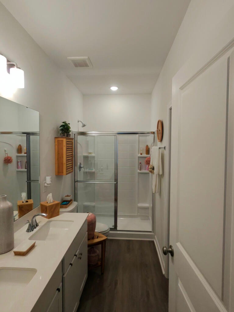 A freshly cleaned bathroom with dual sinks, a walk-in shower, and soft wood flooring. White towels, warm lighting, and subtle decor create a calm, cared-for atmosphere. The space feels organized, inviting, and professionally maintained.